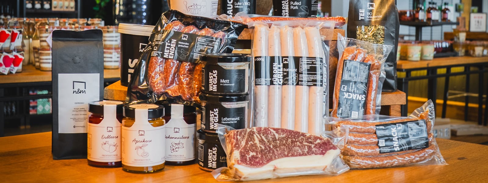 Eine Auswahl an Fleisch und Beilagen, darunter Würste, Steak und Saucen, präsentiert auf einem Holztisch in einer Ladenumgebung.