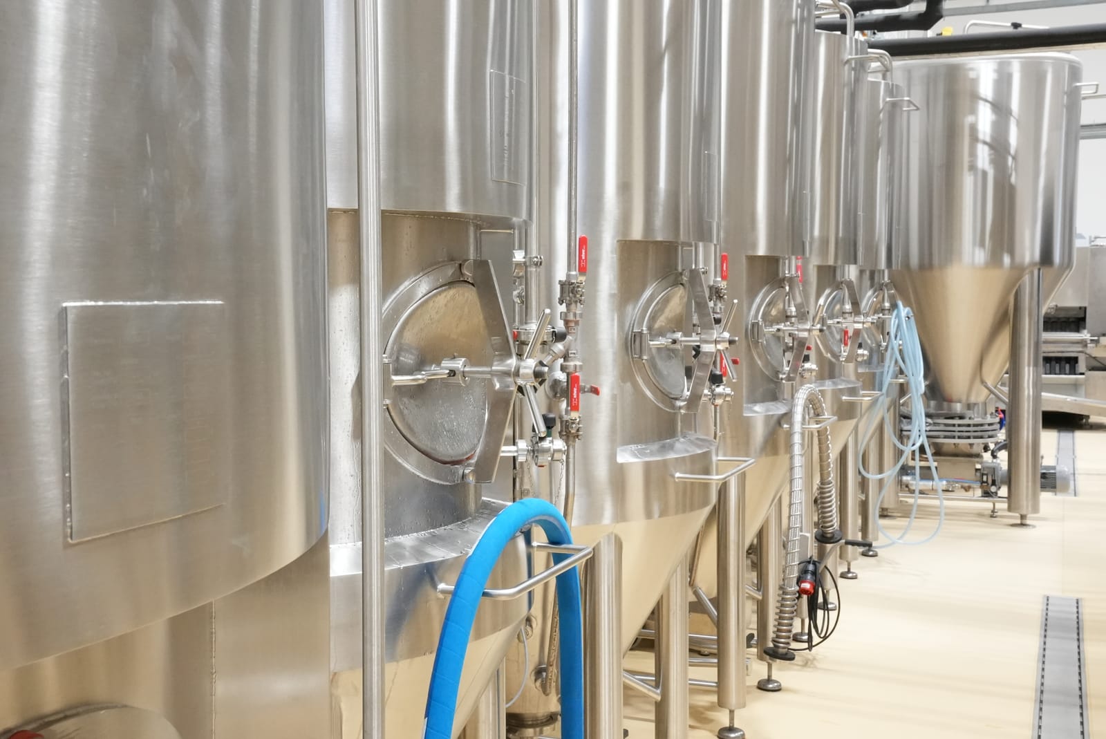 Reihe von Edelstahl-Gärtanks in einer Brauerei, ausgestattet mit Ventilen und Schläuchen für die Bierproduktion.