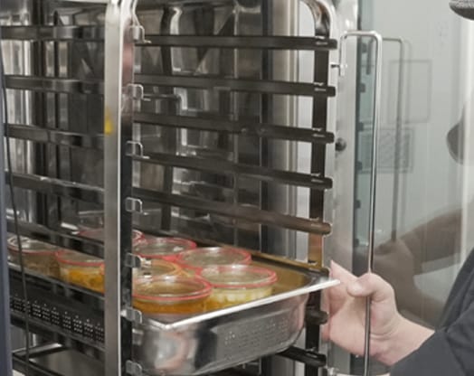 Eine Person stellt verschlossene Gläser mit buntem Inhalt auf ein Metallgestell in einem gewerblichen Ofen.