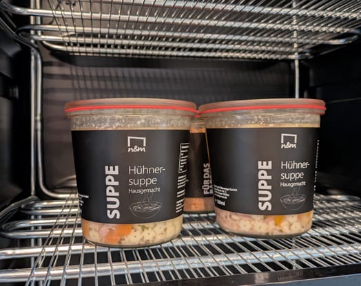 Zwei Gläser hausgemachte Hühnersuppe mit der Aufschrift „Suppe“ auf einem Metallgestell im Backofen. Die Gläser haben rote Deckel und schwarze Etiketten.