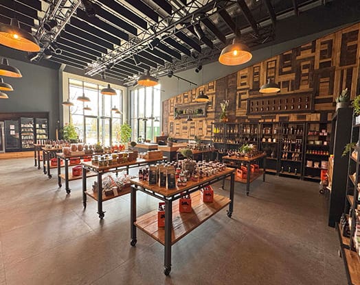 Ein geräumiger Laden mit Holzregalen und Tischen, auf denen verschiedene Produkte ausgestellt sind. Warme Beleuchtung und große Fenster erhellen den Raum.