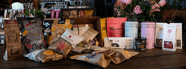 Eine Präsentation verschiedener Gourmet-Snackpakete auf einem Holztisch, umgeben von einer Vase mit rosa Blumen in einer gemütlichen Atmosphäre.