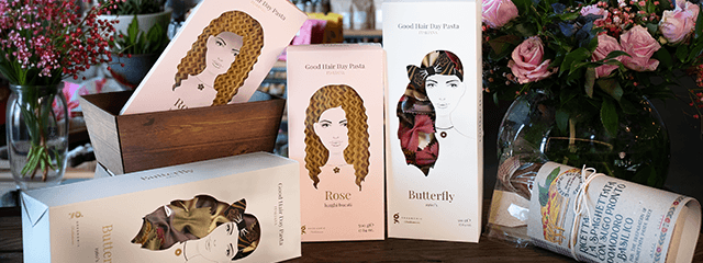 Verschiedene verpackte Teigwaren präsentiert zusammen mit Blumenarrangements; die Verpackungen zeigen Illustrationen von Frauen und anstelle der Haare sieht man die Nudelsorten durch eine transparente Folie, die Packungen sind mit den Sortennamen „Rose“ und „Butterfly“ beschriftet.