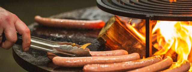 Würste, die über offener Flamme gegrillt werden, während eine Hand mit einer Zange sie auf einem runden Metallgrill wendet.