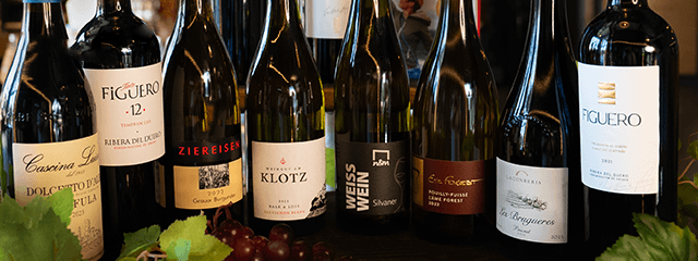 Eine Auswahl von acht Weinflaschen, beschriftet mit verschiedenen Namen und Logos, arrangiert auf einer dunklen Oberfläche, daneben Weinblätter und Trauben.