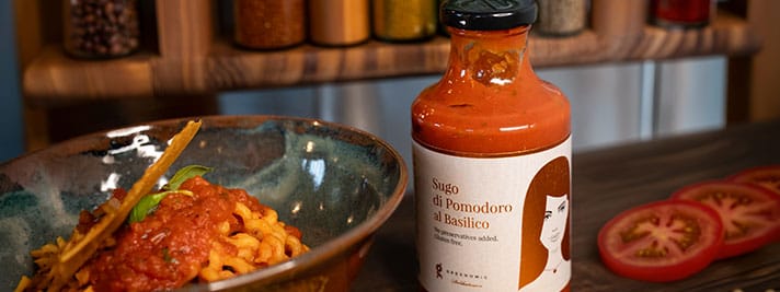 Eine Schüssel Pasta mit Tomatensauce und ein Glas mit der Aufschrift „Sugo di Pomodoro al Basilico“. Auf dem Tisch liegen geschnittene Tomaten, im Hintergrund sind Gewürze zu sehen.