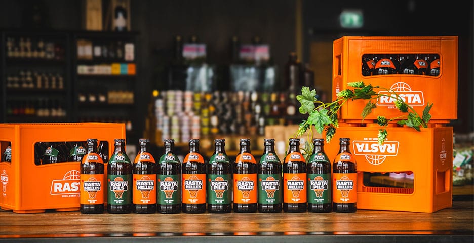 Reihen von RASTA-Getränkeflaschen auf einer Theke, flankiert von Orangen-Kisten, mit einem Blattzweig als Dekoration. Im Hintergrund sind Regale zu sehen.