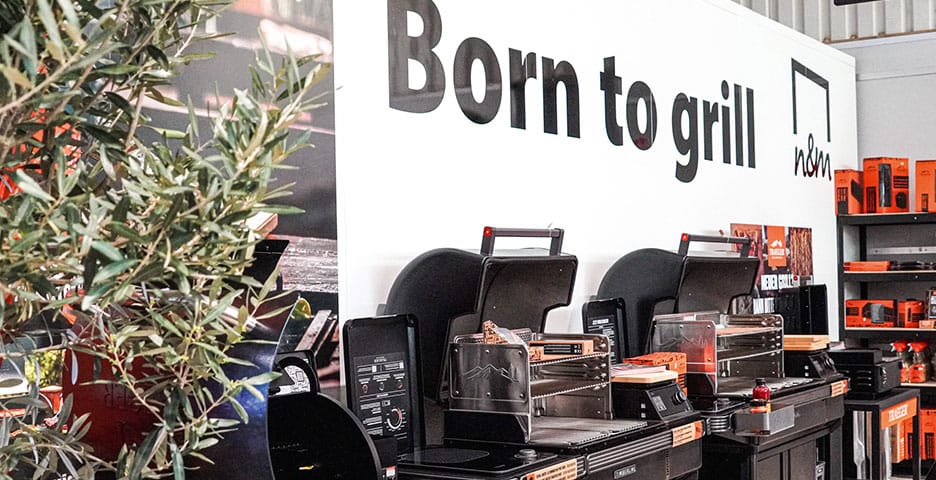 Ausstellung von Outdoor-Grills mit verschiedenen Grillmodellen, einem Schild mit der Aufschrift „Born to grill“, Regalen mit Zubehör und einer Topfpflanze links daneben.