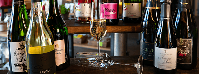 Eine Auswahl verschiedener Schaumweinflaschen, die auf Holzregalen präsentiert werden, mit einem gefüllten Champagnerglas und einem leeren Glas im Vordergrund.