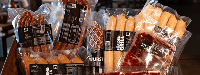 Eine Auswahl verpackter Wurstwaren, darunter Würste und geschnittener Schinken, aufrecht präsentiert.