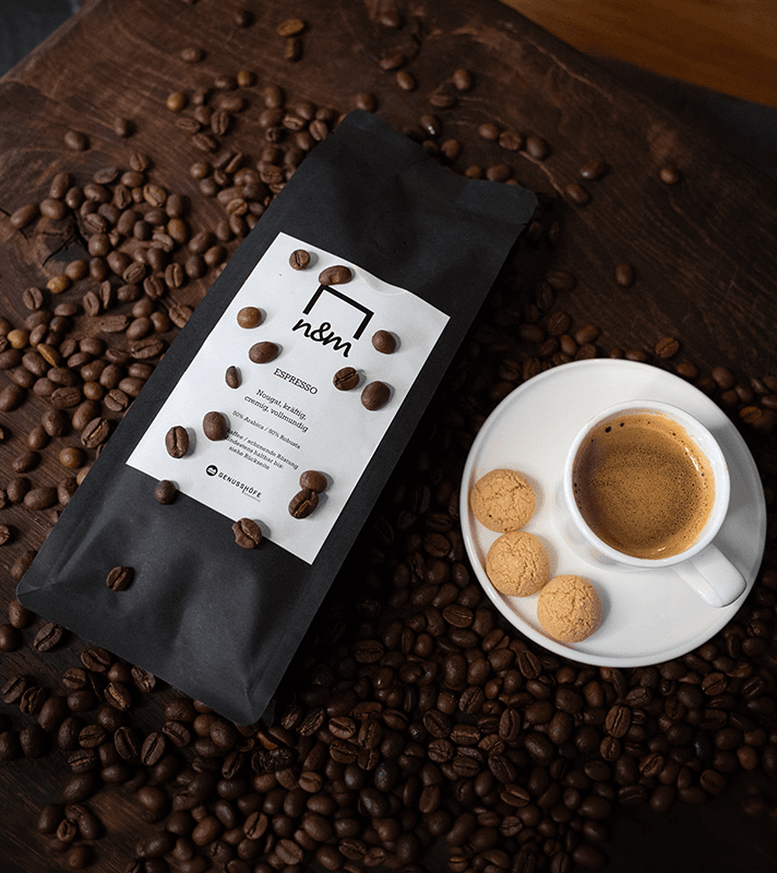 Schwarze Kaffeebohnen-Tüte mit weißem Etikett, umgeben von Kaffeebohnen, daneben eine weiße Tasse Espresso mit drei Keksen auf einem weißen Unterteller.