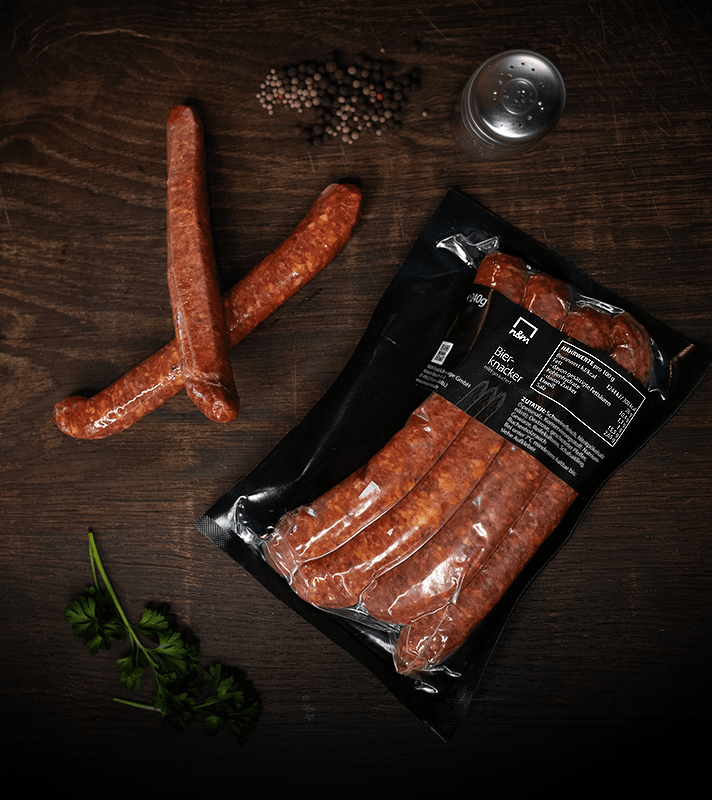 Vier rotes Würstchen in einer schwarzen Verpackung, daneben zwei weitere Würstchen, Pfefferstreuer, frische Petersilie und ein Salzstreuer auf dunklem Holz.