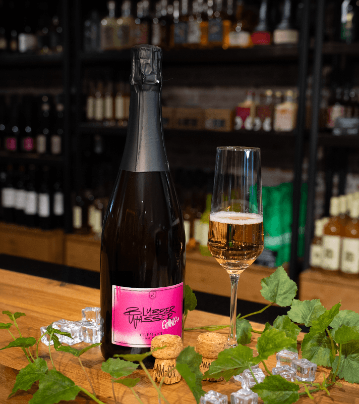 Sektflasche mit schwarzem Verschluss und pinkem Etikett, das den Namen "Blubber Wasted Gang" und "Crement Rose Brut" zeigt, steht zusammen mit einem befüllten Sektglas auf einem Holztisch mit Getränkeregal im Hintergrund.