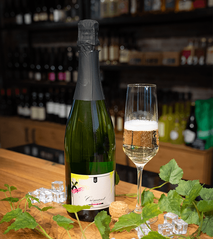 Sektflasche mit Schaumwein, geöffnet, neben einem Glashalter mit Sektglas, auf einem Holzbar-Tresen mit Eiswürfeln und grünen Weinblättern, im Hintergrund Weinregale.