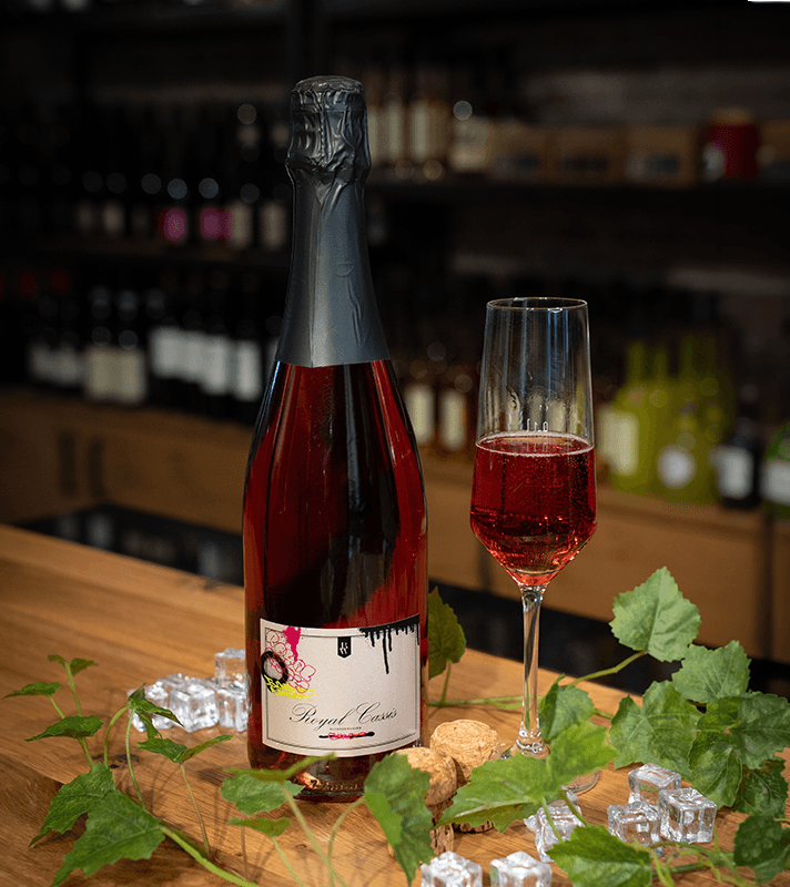 Flasche mit rotem Sekt und ein Glas mit rotem Getränk auf Holzbar, umgeben von Eiswürfeln, Korken und grünen Weinblättern, im Hintergrund Weinregale.