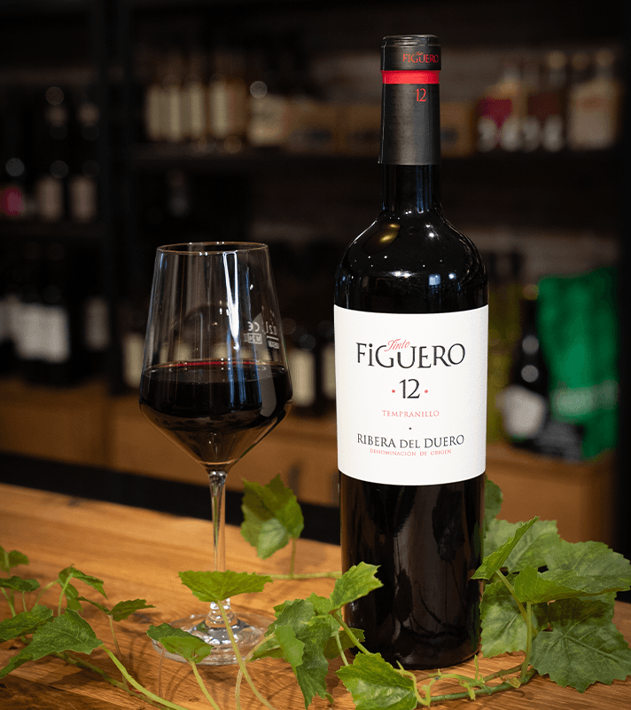 Flasche Rotwein "Figuero 12 Tempranillo" neben einem Weinglas mit Rotwein, auf einem Holztablett mit grünen Weinblättern, im Hintergrund eine Weinregal.