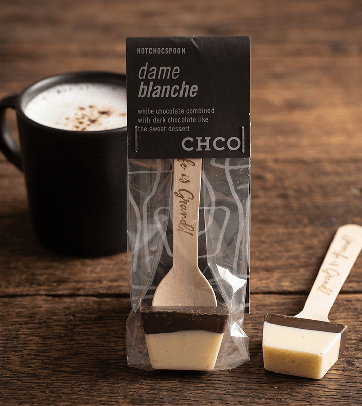 Ein Schokoladen-Eis am Stiel mit weißer und dunkler Schokolade, verpackt in transparentem Kunststoff, auf einem Holztablett neben einer schwarzen Tasse mit Kaffee.
