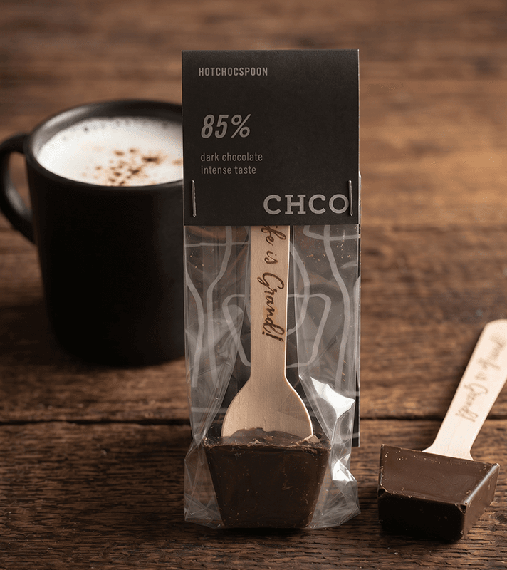 Schokoladenstück mit Holzlöffel, auf dem steht "It's Gourmet!", in transparentem Beutel, dunkle Schokolade mit 85% Kakao, für intensiven Schokoladengeschmack, vor einer Tasse Kaffee auf Holz.