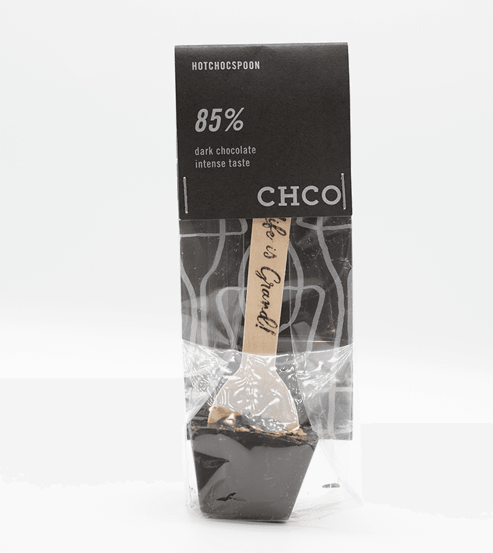 Schwarze Schokoladentafel mit einem Holzlöffel, der mit "handmade" beschriftet ist, in einer transparenten Verpackung. Oberes Etikett mit "85% dunkle Schokolade, intensiver Geschmack".
