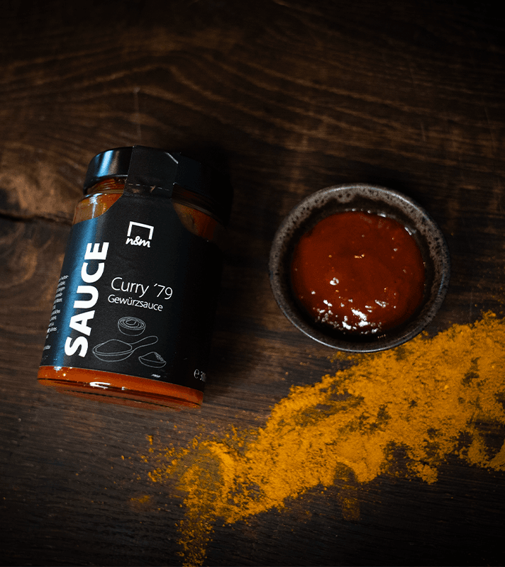 Ein Glas mit Curry-Gewürzsauce neben einem kleinen Schälchen mit Sauce und einer Gewürzspur auf dunklem Holz.