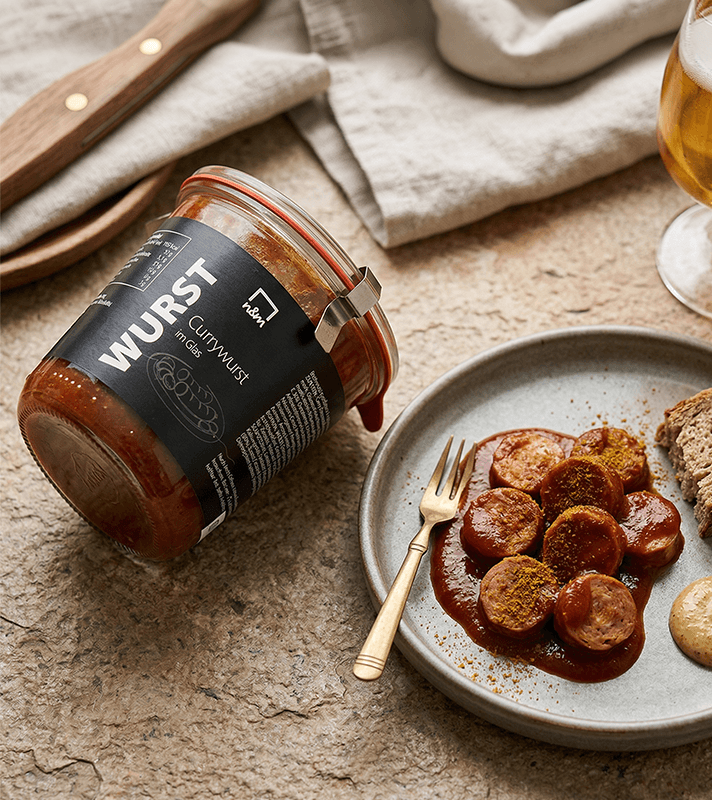 Glas mit Currywurst in Sauce, mit schwarzer Etikette, weißem Text und einem kleinen Illustrationssymbol einer Currywurst auf der Vorderseite.