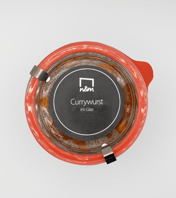 Glasbehälter mit Currywurst im Glas, abgedeckt mit einem roten Deckel und Metallverschlüssen, schwarzes Etikett mit weißen Schriftzug.