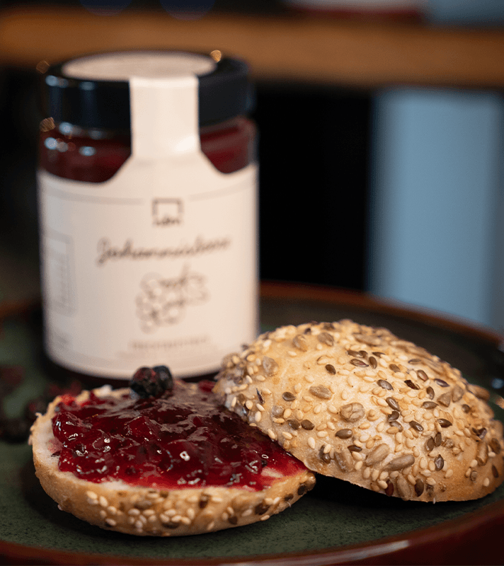 Ein belegtes Brötchen mit roter Fruchtmarmelade, daneben ein weiteres Brötchen mit Saaten, auf einem dunklen Teller, im Hintergrund eine Marmeladenglas.