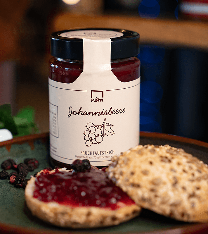 Ein belegtes Brötchen mit roter Fruchtmarmelade, daneben die obere Brötchenhälfte mit Saaten, auf einem dunklen Teller, im Hintergrund eine Marmeladenglas.