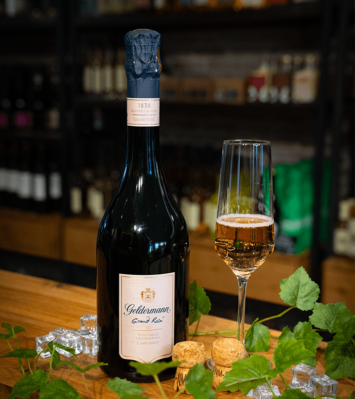 Flasche mit Rosé-Sekt, Glas mit Rosé-Sekt, Korken, Eiswürfel, Weinblattdekorationen auf Holzoberfläche.