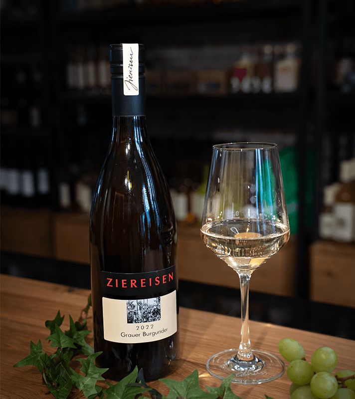 Eine Flasche Weißwein mit schwarzem Etikett und weißem Label, daneben ein Weinglas mit Weißwein, grüne Weintrauben und grüne Weinblätter auf einem Holztisch.