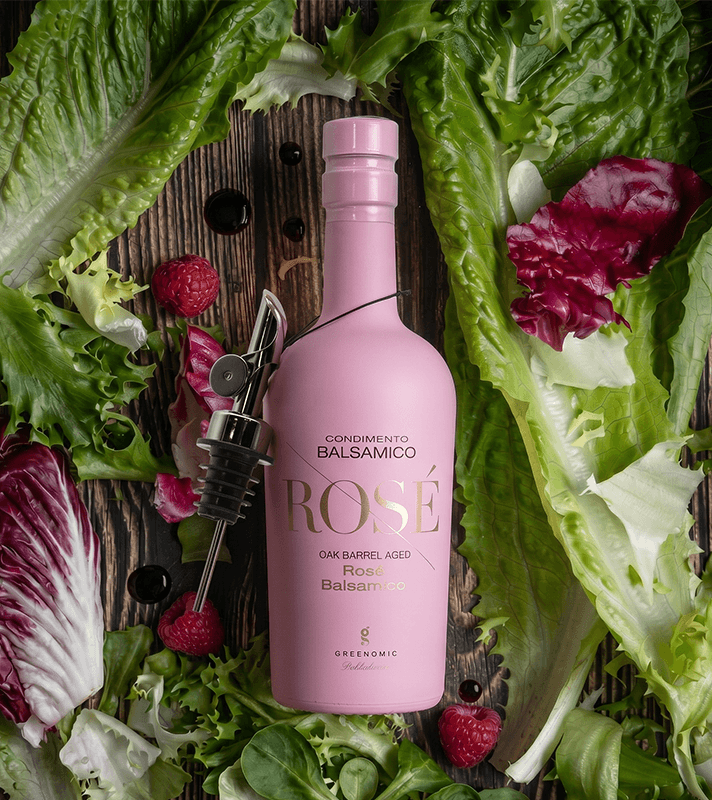 Rosa Balsamico Wein mit rosa Flasche, umgeben von grünen und roten Salatblättern, Himbeeren und schwarzen Beeren, mit einem Weinverschluss.