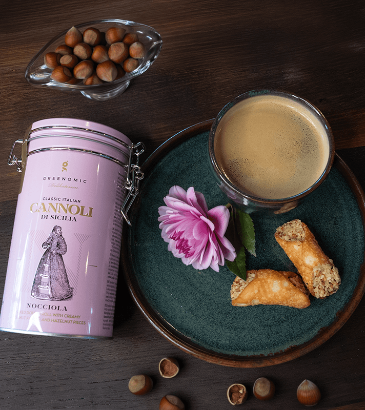 Eine Helllila farbene Dose Cannoli liegt auf einem dunklen Tisch neben einem dunklen Teller mit einem Anrichtungsbeispiel der Cannoli