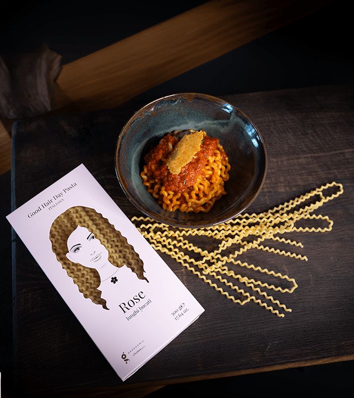 Eine Schüssel mit gekochten Nudeln in Tomatensauce, garniert mit einer Scheibe Parmesan, auf einem dunklen Holzbrett. Daneben liegen ungekochte Pasta und eine Illustration einer Frau mit lockigem Haar.