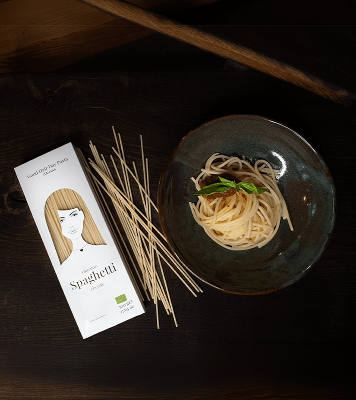 Schüssel mit gekochten Spaghetti, frischer Basilikum und Tomaten, daneben trockene Spaghetti und eine Verpackung mit Illustration einer Frau mit langen Haaren.