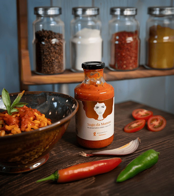 Flasche mit roter Soße, umgeben von Tomatenscheiben, Chili, Zwiebel und einem Teller mit Pasta und Basilikum, im Hintergrund Gewürzgläser auf Holzregal.