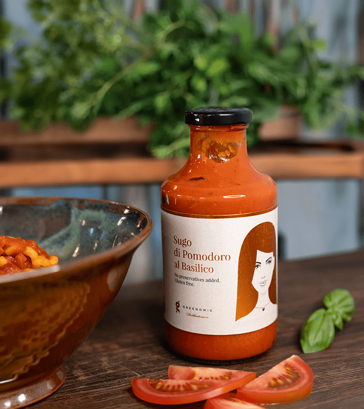 Flasche mit Tomatensugo im Glas, Etikett mit Frauenillustration, daneben Tomatenscheiben, im Hintergrund grüne Pflanzen und eine Schüssel mit Pasta.