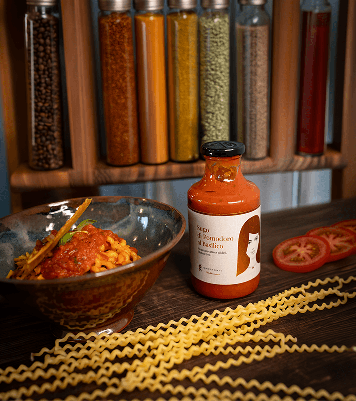 Flasche mit Tomatensugo im Glas, Etikett mit Frauenillustration, daneben Tomatenscheiben, im Hintergrund grüne Pflanzen und eine Schüssel mit Pasta.