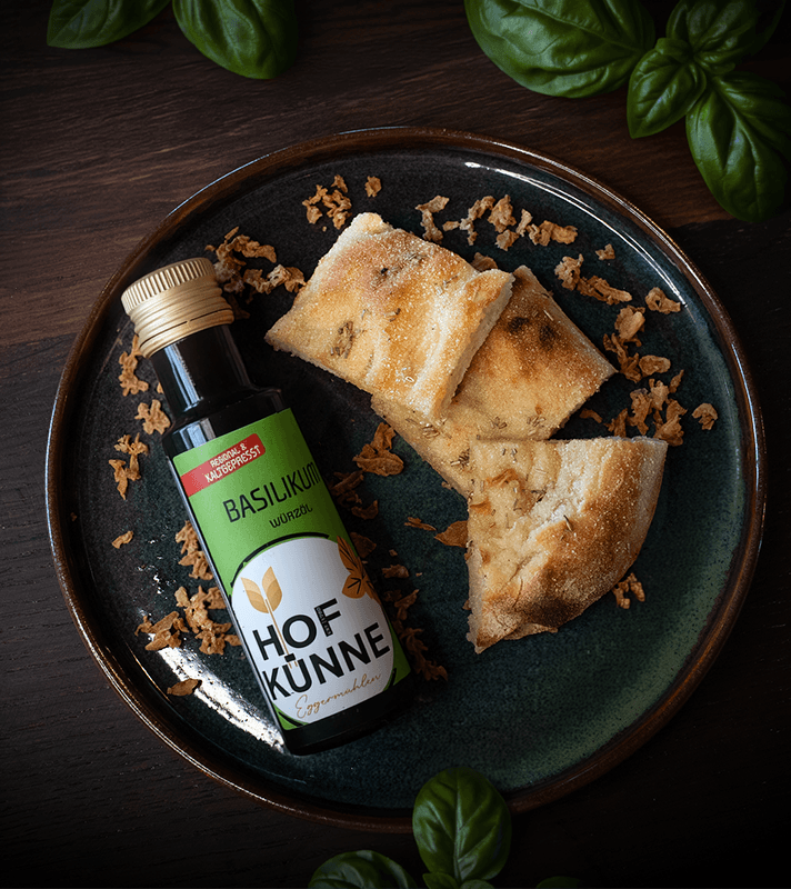 Eine Flasche Basilikum Würzöl liegt auf einem dunklen Teller mit angerichteten Brotstücken, der Teller steht auf einer dunkelbraunen Arbeitsfläche