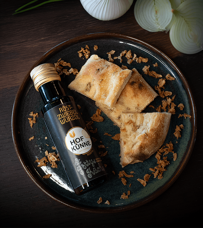 Eine Flasche Röstzwiebel Würzöl liegt auf einem dunklen Teller mit angerichteten Brotstücken, der Teller steht auf einer dunkelbraunen Arbeitsfläche