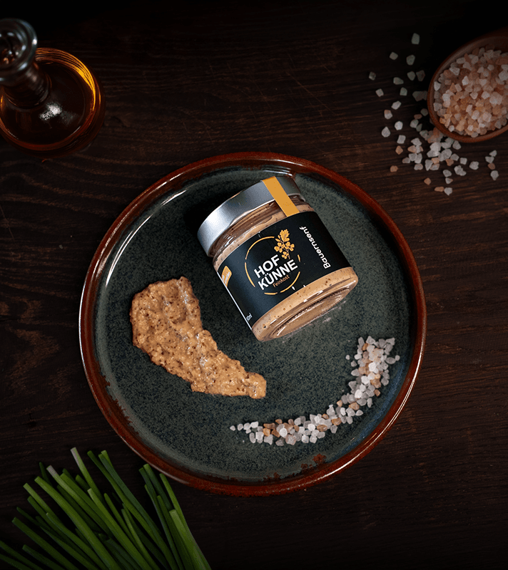 Glas mit Senf auf dunklem Teller, daneben Salz, Pfeffer und frische Schnittlauchstangen auf dunklem Holztisch.