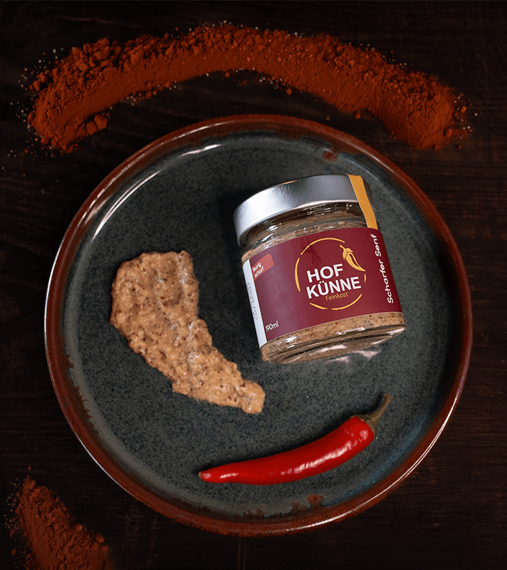 Glas mit Fleischwürstchenpaste, rote Chili, Paprikapulver im Hintergrund, auf dunklem Holzuntergrund.