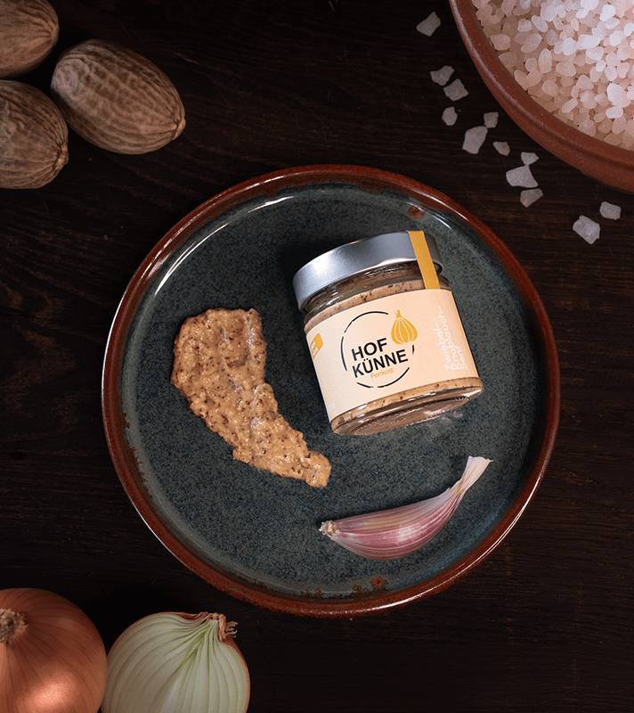 Kleiner Glas mit Gewürzpaste, daneben eine Knoblauchzehe, auf einem blauen Teller. Im Hintergrund Salz in einer Schale und ganze Muscheln.