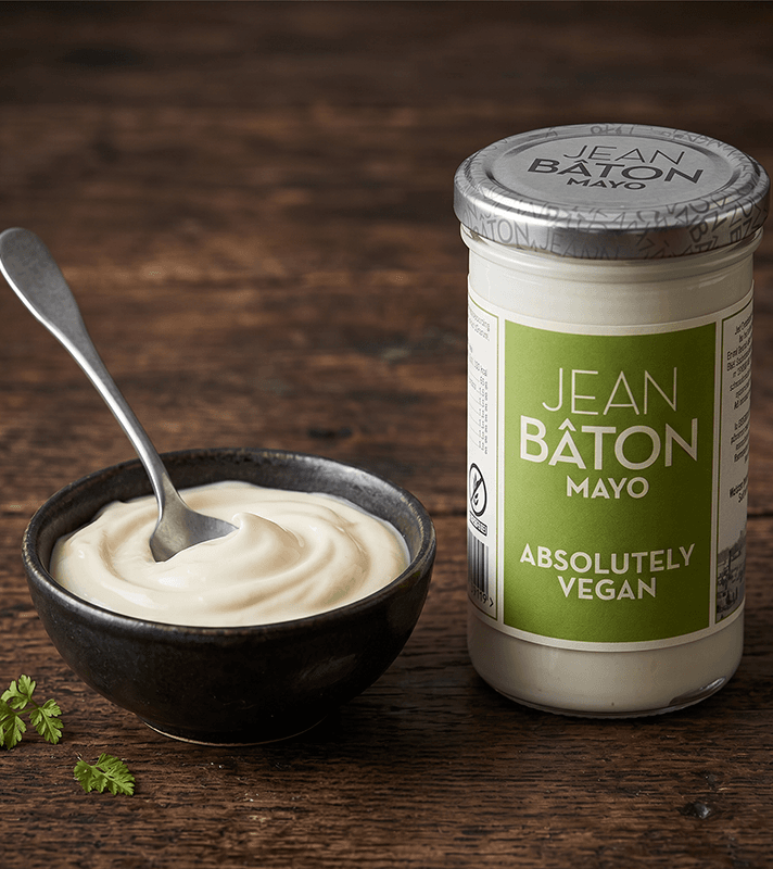 Vegane Mayonnaise in einem schwarzen Schälchen mit Löffel, daneben ein Glas mit grünem Etikett, auf dem "Jean Bâton Mayo Absolutely Vegan" steht, auf dunklem Holzuntergrund.