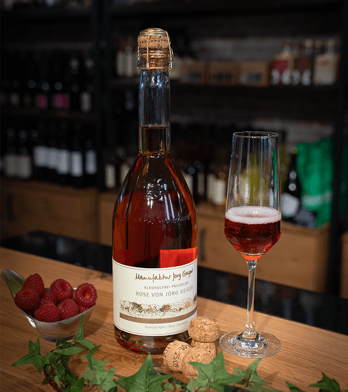Alkoholfreier Rosé-Sekt mit rotem Fruchtgeschmack, serviert in einem Sektglas neben frischen Himbeeren und Korken auf einem Holztablett.