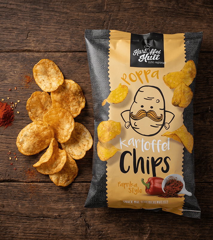Bunte Kartoffelchips in Paprika-Style auf einer Holzoberfläche, neben einer geöffneten Packung mit einem lustigen Kartoffel-Charakter.