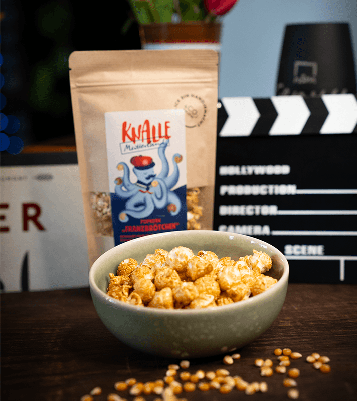 Beutel mit Popcorn "Franzbrötchen" von Knalle, mit knusprigem, karamellisiertem Popcorn, neben einer Schüssel mit Popcorn und einer Maiskolbe auf dunklem Holz.