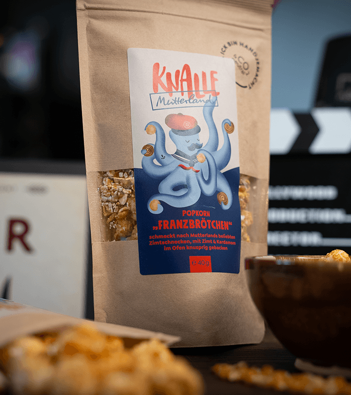 Beutel mit Popcorn "Franzbrötchen" von Knalle, mit knusprigem, karamellisiertem Popcorn, neben einer Schüssel mit Popcorn und einer Maiskolbe auf dunklem Holz.