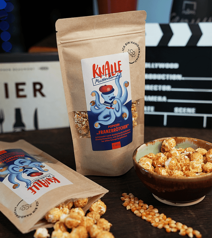 Beutel mit Popcorn "Franzbrötchen" von Knalle, mit knusprigem, karamellisiertem Popcorn, neben einer Schüssel mit Popcorn und einer Maiskolbe auf dunklem Holz.