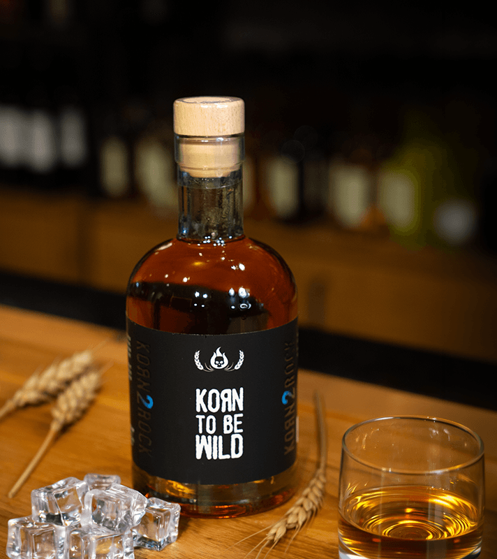 Kleiner Glas mit goldener Spirituose, daneben eine Flasche mit schwarzem Etikett, auf dem "KOAN TO BE WILD" steht, und zwei Weizenähren auf einem Holztablett.