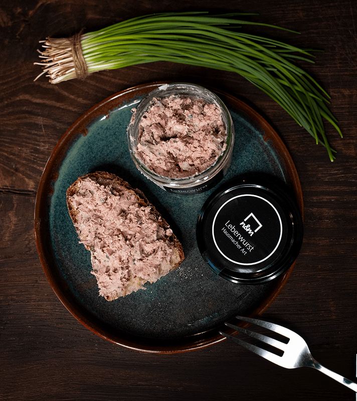 Auf einem braunen Teller liegt eine Scheibe Brot mit Leberwurst. Daneben ein offener Glasbehälter mit Leberwurst und ein schwarzer Deckel. Im Hintergrund liegt frische Petersilie.